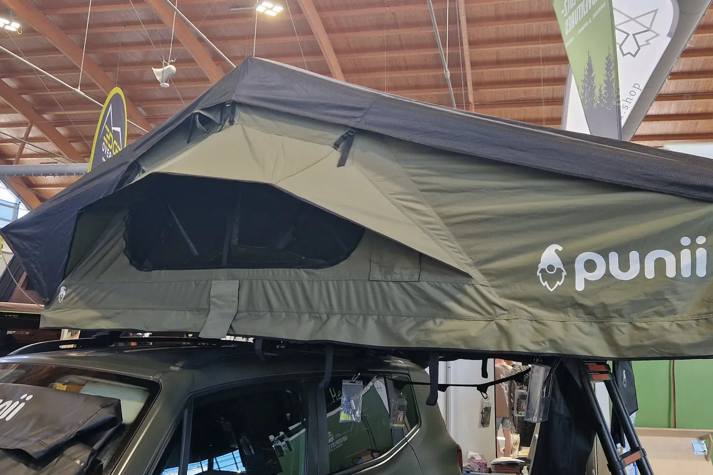 Olivgruenes punii Softshell-Klappdachzelt seitlich aufgeklappt auf einem Fahrzeug am Messestand