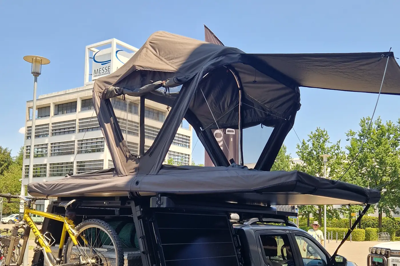 Nissan Navara Pickup mit ROOF SPACE Klappdachzelt, Aluminium-Plattform und ausgeklappter 270-Grad-Markise auf einer Messe in Friedrichshafen