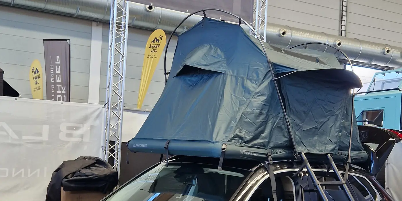 Dunkelblaues Softshell-Klappdachzelt mit Bogengestaenge und Leiter auf einem SUV in der Messehalle